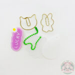 Dos sujetadores o señaladores de hojas de color dorado con forma de gato y conejo. Broche rosa con la palabra Lucky. Dos dijes blancos con forma de gota y flor con cadena para colgarlos.