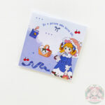 Bloc de notas con estampado cuadriculado celeste y diseño con dibujos, en la parte derecha una chica con vestido azul y en la parte izquierda y central aparecen una cesta de picnic, libro, cereza.